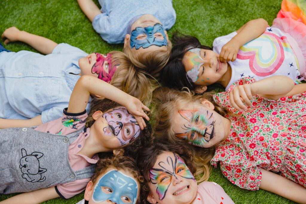 pexels-photo-8385033-8385033 Group of children enjoying an outdoor party with colorful face paint, lying on grass.
