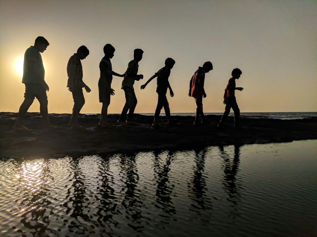 pexels-photo-939700-939700 A group of children walk along the shoreline, silhouetted against a sunset, reflecting in the water.